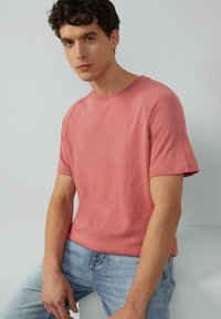 Koszulka z krótkim rękawem, okrągłym dekoltem w jasnym koralowym kolorze z delikatną teksturą. Zestawiona z jasnoniebieskimi dżinsami. Model siedzi na szarym tle.