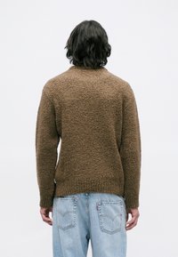 Persona con cabello oscuro hasta los hombros, vistiendo un suéter marrón texturizado y jeans de mezclilla azul claro, de espaldas a la cámara.