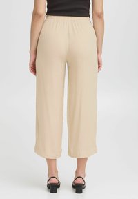 Beige wijde broek met een gekreukte textuur, een elastische tailleband en een cropped lengte. Draagt met zwarte sandalen met blokhak.