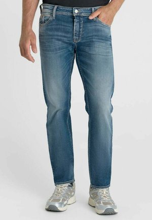 Man draagt vervaagde blauwe jeans en wit-grijze sneakers, staand met één hand in de zak tegen een effen achtergrond.