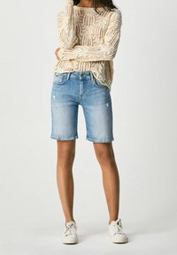 Bermudas de mezclilla azul claro con un acabado ligeramente desgastado, combinadas con un suéter de punto texturizado en color crema y zapatillas blancas.