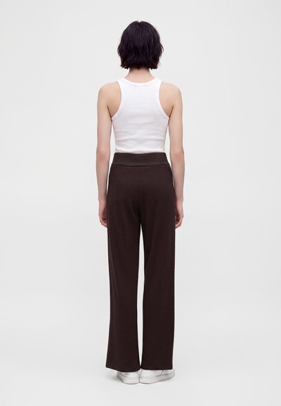 Personne aux cheveux courts et foncés portant un haut blanc sans manches, un pantalon large foncé et des baskets blanches vue de dos sur un fond uni.