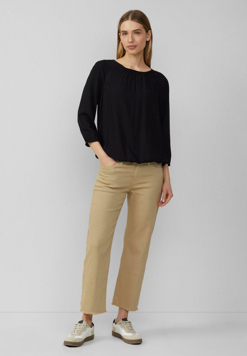 Zwarte blouse met plooien bij de halslijn, gecombineerd met lichtbruine cropped pantalons. Model draagt witte en grijze sneakers met veters.