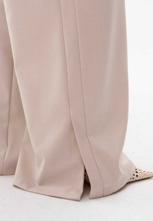 Jasno beżowe spodnie w szerokim kroju o miękkiej fakturze i delikatnych detalach plis, lekko rozszerzane na końcach, zestawione z plecionymi beżowymi butami.