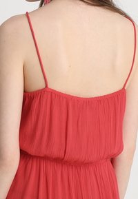 Robe rouge légère sans manches avec de fines bretelles et un design texturé et fluide. Ceinture élastique et détail de tissu froncé en haut.