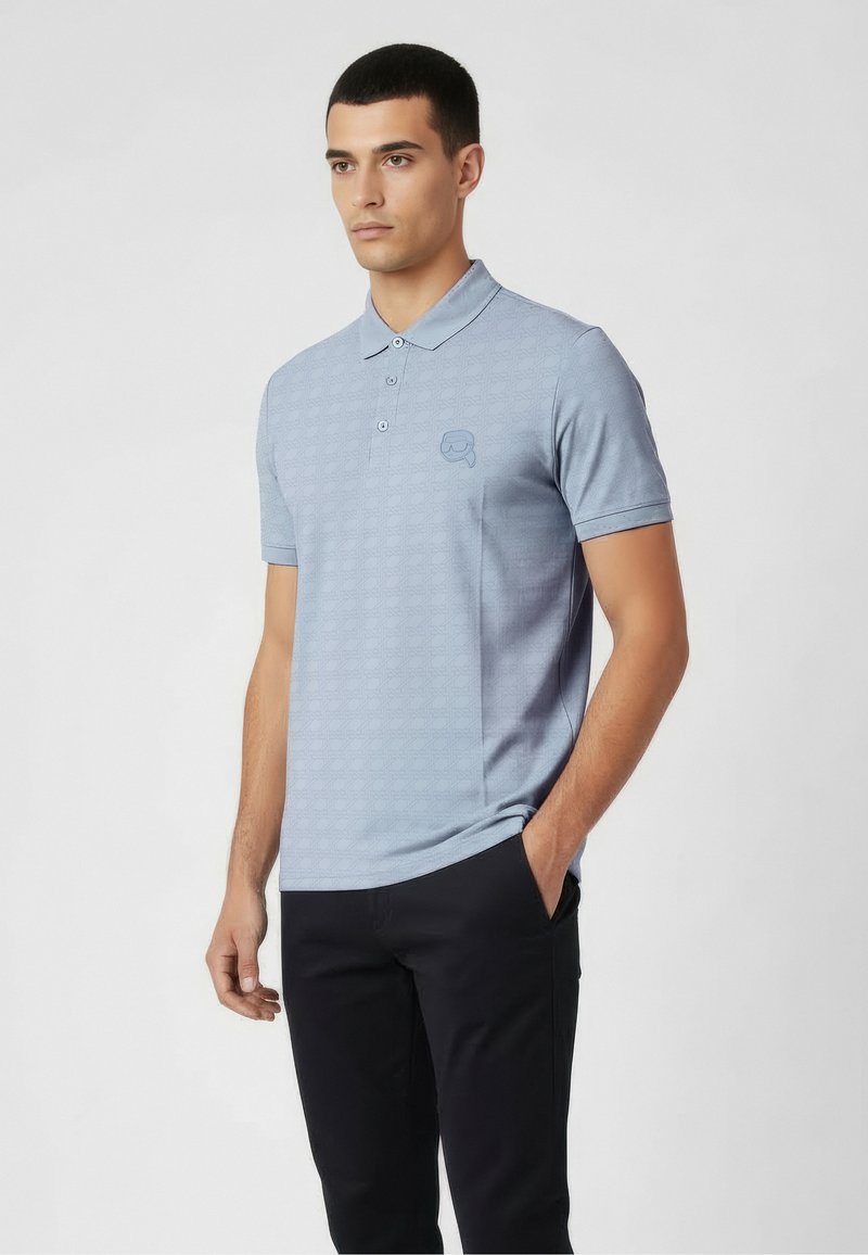 Polo de color azul claro con textura sutil de patrón, tapeta de dos botones y un pequeño logo en el pecho izquierdo. Mangas cortas, ajuste regular.