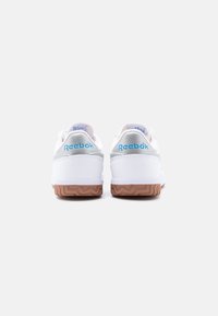 Ζευγάρι λευκά αθλητικά παπούτσια Reebok, εμφανιζόμενα από πίσω με μπλε λογότυπο, γκρι λεπτομέρεια στη φτέρνα και σόλες από καουτσούκ θυμίσματος καραμέλας σε απλό φόντο.