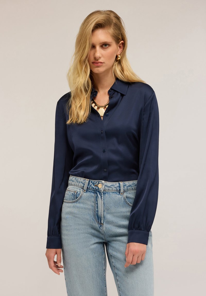 Donna bionda che indossa una camicetta blu satinata con bottoni, jeans a vita alta di colore blu chiaro, collana con catena dorata e orecchini a cerchio dorati.