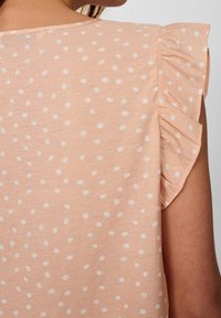 Blusa pesca con pois bianchi, dotata di maniche arricciate e una texture liscia. La vista posteriore mostra un collo rotondo e una leggera lucentezza.