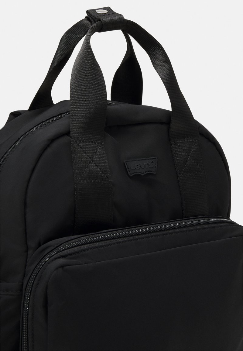 Sort rygsæk lavet af slidstærkt stof med polstrede stropper, der har en frontlomme med lynlås og et lille logo-patch. Glat tekstur med diskret syning.
