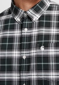 Vihreä-musta ruudullinen nappipaita, jossa valkoiset napit, kaulus ja pieni logo taskussa. Kangas on kudottu teksturoidulla kuviolla.