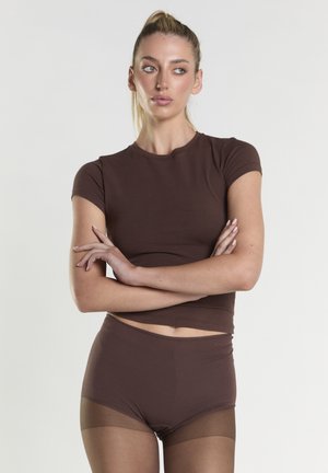 Kvinde med blond hår i en hestehale har en brun top med korte ærmer og matchende shorts på sammen med gennemsigtige brune strømpebukser, armene krydset, ser mod højre.
