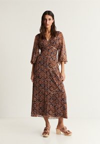 Bruine maxi-jurk met patroon, een V-hals, uitlopende mouwen en een luchtige silhouette. Gecombineerd met bruine hoge klompen met een strikdetail.