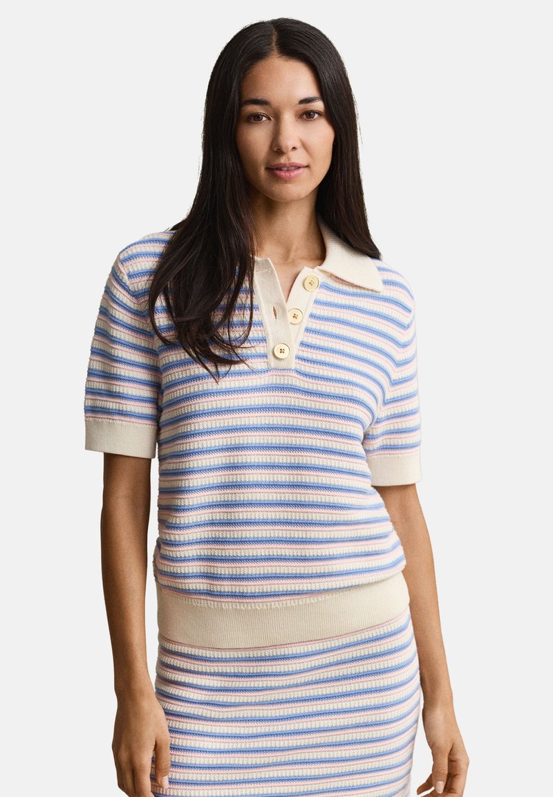 Γυναίκα με μακριά σκούρα μαλλιά που φοράει μπλε και μπεζ ριγέ πλεκτή μπλούζα με κοντά μανίκια και κουμπιά, καθώς και φούστα που ταιριάζει.