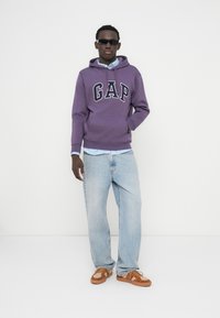 Sweat à capuche violet avec le logo « GAP », chemise à carreaux bleu clair en dessous, jean bleu clair, et chaussures marron à enfiler avec un motif. Lunettes de soleil noires.