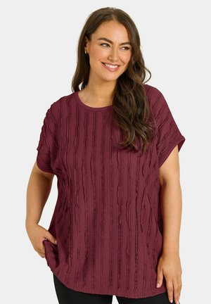 Vrouw met lang bruin haar die een gestructureerde bordeauxkleurige top met korte mouwen draagt, glimlachend en zijwaarts kijkend met één hand in haar zak.
