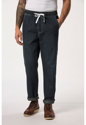 Pantalones jogger de denim azul oscuro con cintura ajustable mediante cordón, costuras blancas y puños enrollados. Botas marrones con cordones complementan el conjunto.