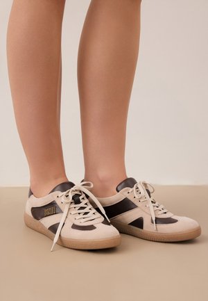 Benen met beige en donkerbruine vetersneakers met rubberzolen op een neutrale vloer en achtergrond.