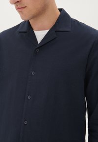 Tengerészkék vászonting kerekített gallérral, elülső gombokkal és texturált anyaggal. A dizájn hangsúlyozza a laza szabást és az egyszerűséget.