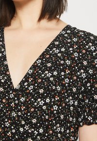 Femme portant une robe noire à col en V avec un petit motif floral blanc, orange et vert, et des manches courtes bouffantes.