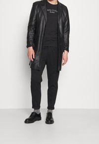 Abrigo de cuero negro, camiseta negra con texto, pantalones negros y zapatos negros de cordones. Calcetines grises y con los puños de los pantalones enrollados.