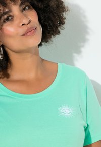 Mátově zelené bavlněné tričko s kulatým výstřihem a bílým vyšívaným motivem oka s paprsky na levé straně. Hladká texturovaná látka.