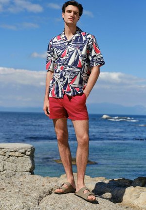 Mladý muž stojící na skalnatém pobřeží, oblečený v krátkém tričku s potiskem plachetnice, červených šortkách a béžových sandálech s oceánem v pozadí.