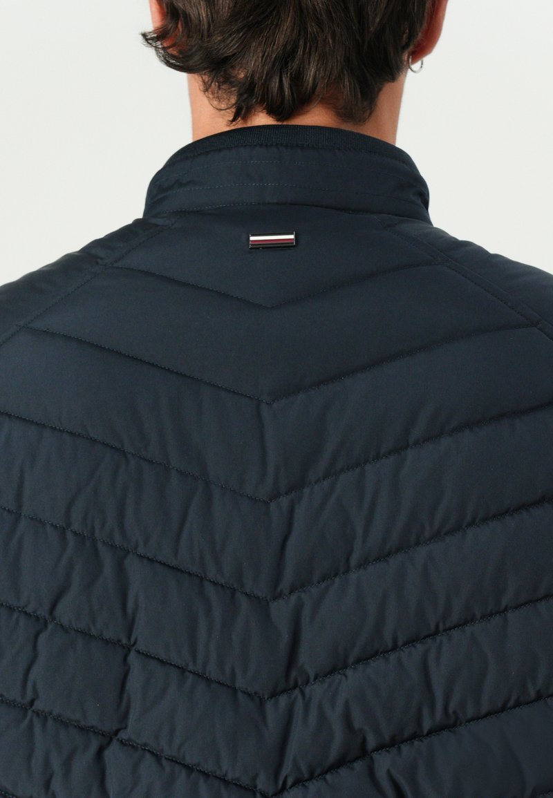 Granatowa kamizelka pikowana w wzór chevron, z małym metalicznym logo na kołnierzu z tyłu oraz gładką teksturą na zewnątrz.