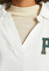 Bílá polo košile s výstřihem do V, přes béžový rolák. Má grafiku zeleného písmene 'P' a texturovanou tkaninu.
