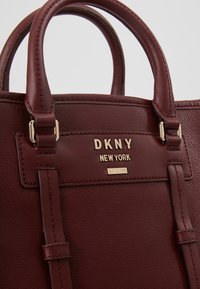 Sac à main en cuir bordeaux avec deux poignées courtes, logo embossé en or et forme rectangulaire ; comporte des accents métalliques dorés.