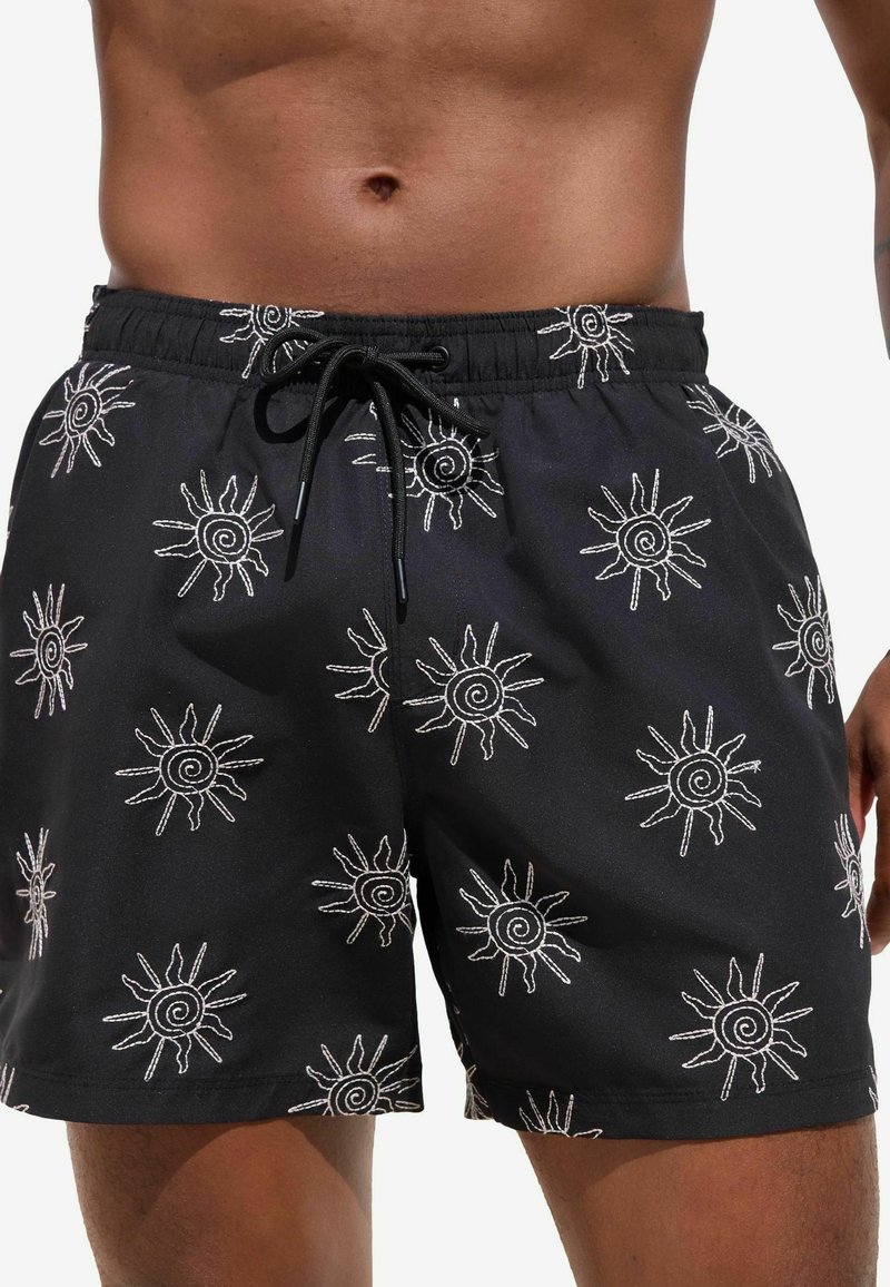 Svarta badshorts med vita spiralmönster av solen, elastisk midja med snörning, lättviktigt tyg och kortare snitt ovanför knäet.