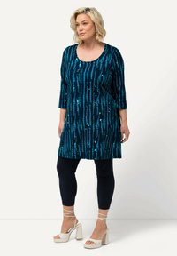 Tunique bleu et teal avec un motif rayé vertical, manches trois-quarts et encolure arrondie, associée à un legging noir et des sandales à talons blocs blanches.