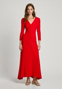 Robe longue rouge à manches longues avec un décolleté en V et des détails froncés à l'avant, en tissu lisse, et une jupe fluide ; associée à des sandales métalliques.