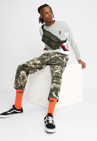 Mala de cintura verde, sweatshirt cinza com logotipo, calças de camuflagem e meias laranjas. Ténis pretos com solas e atacadores brancos. Fundo simples.
