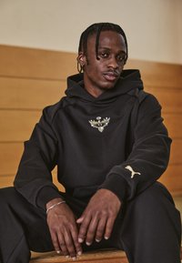 Hanorac negru cu glugă, cu logo brodat al unei vulturi, purtat cu pantaloni asortați. Prezintă logo-ul auriu Puma pe mânecă. Textură de țesătură netedă.