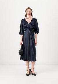 Jūros mėlynos satino suknelė su giliu V formos iškirpimu ir kimono rankovėmis, turinti surištą liemenį ir plazdančią midi ilgio dalį. Juoda rankinė ir aukštakulniai batai.