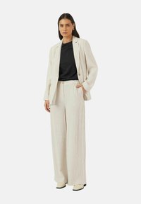 Kremstrimmet, teksturert dress med en skreddersydd blazer og vide bukser, kombinert med en svart topp og lyse sko.