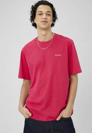 Jeune homme aux cheveux noirs bouclés portant une chemise rouge à manches courtes et une chaîne argentée, mains dans les poches, fond gris neutre.