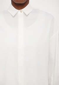 Camicia bianca a maniche lunghe con colletto classico, chiusura frontale a bottoni e tessuto liscio, leggermente trasparente. Presenta un design pulito e minimalista.