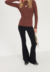 Bruin geribd top met lange mouwen gecombineerd met zwarte geribde uitlopende broek. De outfit wordt aangevuld met een gouden hartketting en zwarte schoenen met metallic accenten.