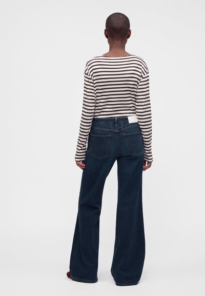 Personne aux cheveux courts portant une chemise rayée à manches longues et un jean large foncé, debout de dos sur un fond uni.