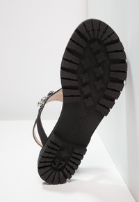 Svart gummisul med djupt mönsterdesign. Har ett dekorativt band med silveraccenter. Storlek markerad som 37. Platt profil, texturerad yta.