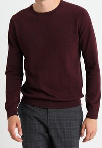 Bordeauxfarbener Rundhalsausschnitt-Pullover aus glatt gestricktem Material, mit langen Ärmeln und schmalem Bund, kombiniert mit grauen Karohosen.