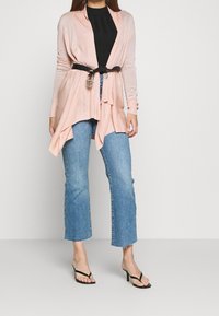 Ljusrosa öppna cardigan med texturerad tyg, bältad vid midjan med ett svart rem. Klagd med blå utsvängdajeans och svarta klacksandaler.