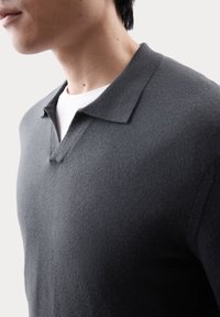Donkergrijze polo trui met een klassieke kraag, gestructureerde stof en een slim fit, gedragen over een wit T-shirt, wat de gelaagde look benadrukt.