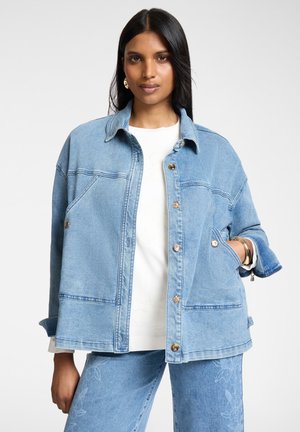 Donna che indossa una giacca di jeans azzurro chiaro e pantaloni abbinati con un motivo floreale sottile, camicia bianca e braccialetti ed orecchini dorati.