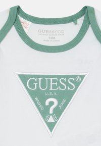 Svijetlosiva organska pamučna bodi s zelenim izvezenim logotipom u obliku trokuta koji sadrži natpis "GUESS U.S.A." i upitnik. Veličina 12M.