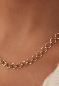 Collier chaîne en or avec des maillons circulaires moyens reposant sur une peau bronzée, avec un bord de vêtement blanc visible en bas à droite.