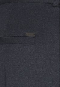Námorně modrá tkanina s texturovaným povrchem, obsahující obdélníkovou kapsu a malou kovovou logovou nášivku. Střihový design s čistými liniemi.