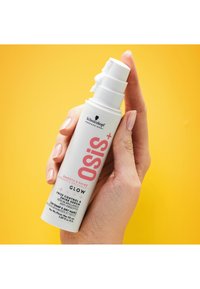 Witte cilindrische fles met roze tekst, vastgehouden door een hand. Voorzien van een pompje en een label waarop staat dat het een glansserum is voor het beheersen van pluis.